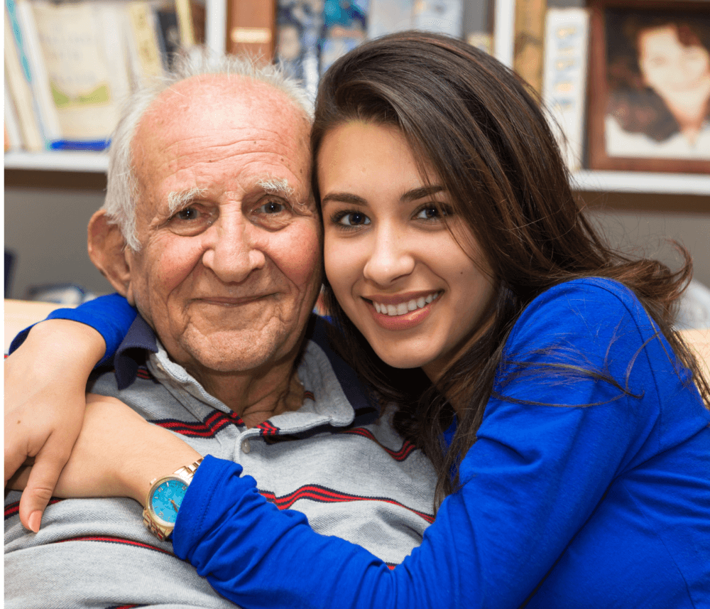 A smiling young woman embraces an elderly man in a cozy room, with books and framed photos in the background. - Home Instead