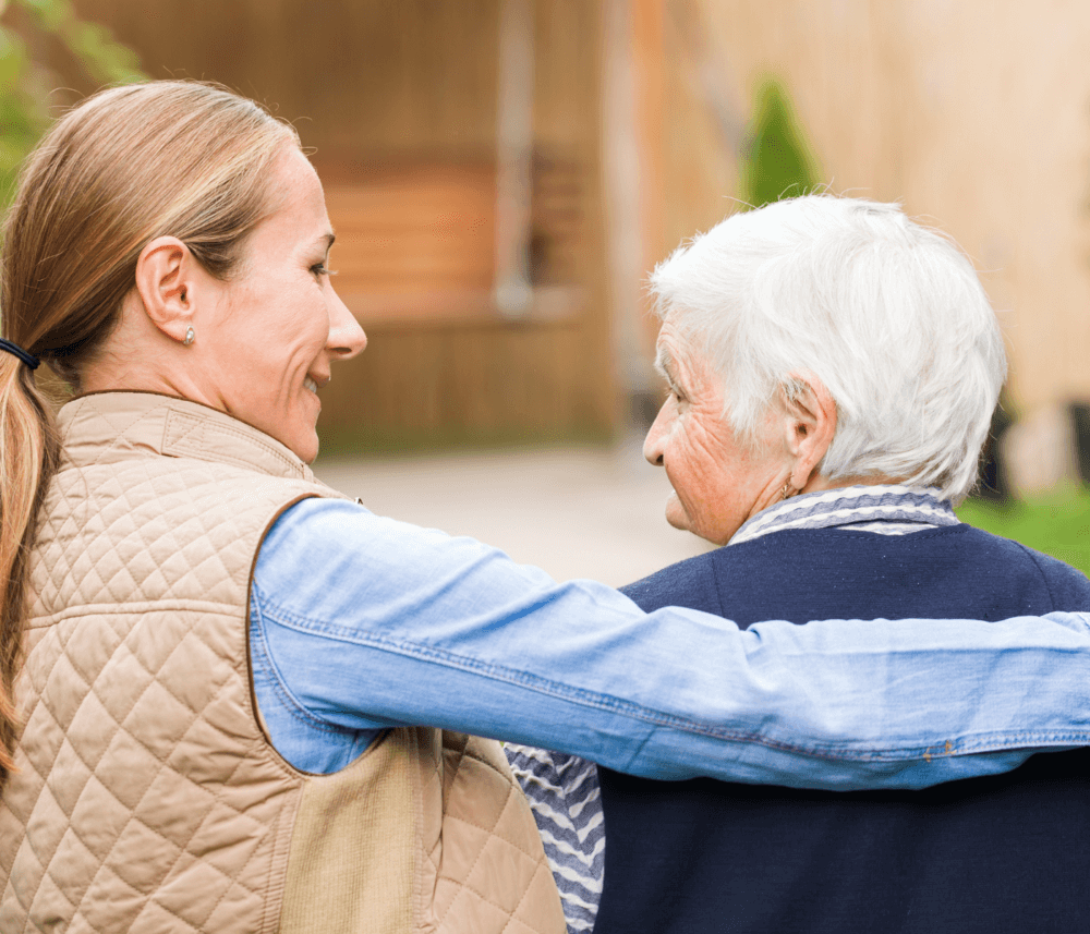 A younger woman with her arm around an older woman, both smiling and looking at each other outdoors. - Home Instead