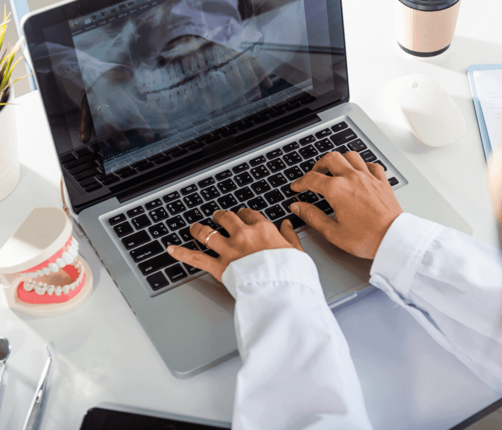 Person in a white coat types on a laptop displaying a dental X-ray, with a dental model, mouse, and cup nearby. - Home Instead