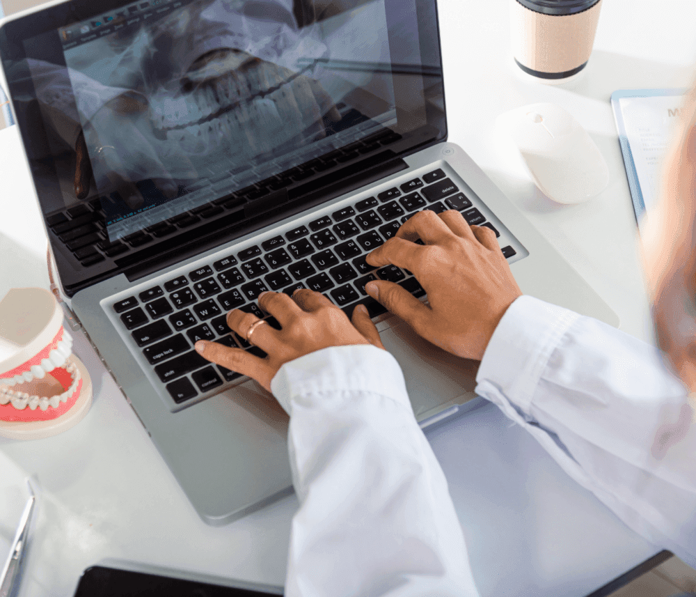 Person typing on a laptop displaying a dental X-ray, with a dental model and a coffee cup nearby on the desk. - Home Instead