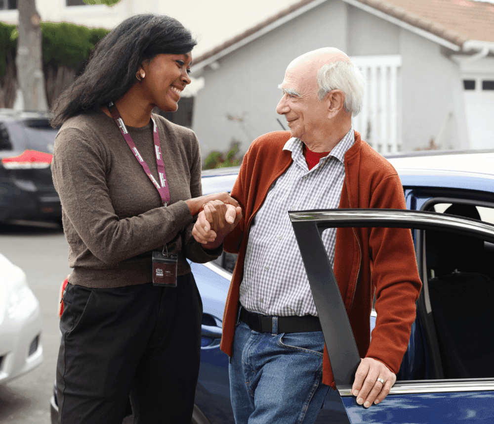 A woman helps an elderly man get out of a car, both smiling and holding hands in a friendly manner. - Home Instead