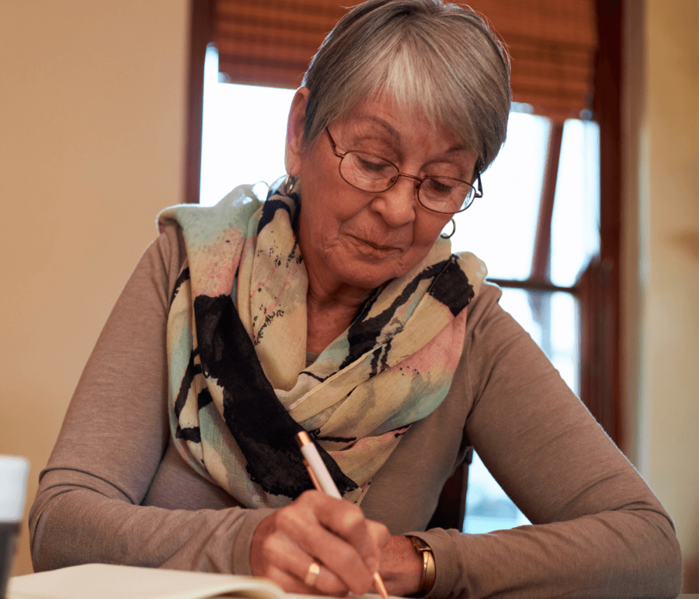 Elderly woman with glasses and a colorful scarf writing in a notebook indoors. - Home Instead