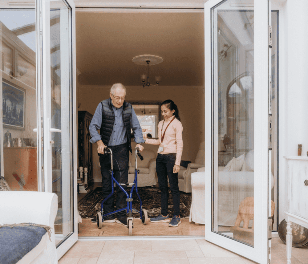 An elderly man using a walker is assisted by a young woman as they step through a doorway into a cozy living room. - Home Instead