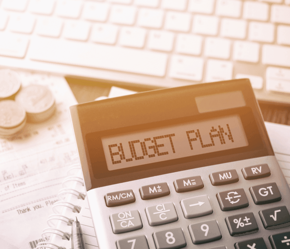 Close-up of a calculator displaying "BUDGET PLAN" with coins, paperwork, and a keyboard in the background. - Home Instead