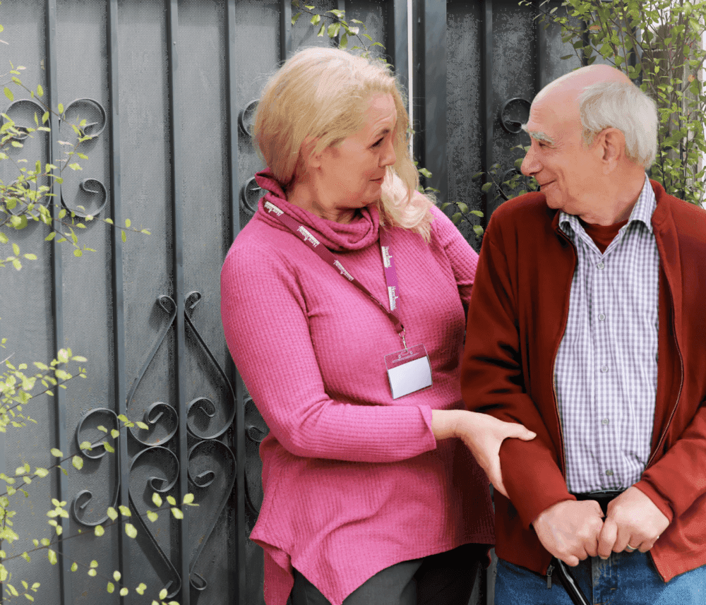 A woman in a pink top talks to an older man in a red sweater near a wrought-iron gate. - Home Instead