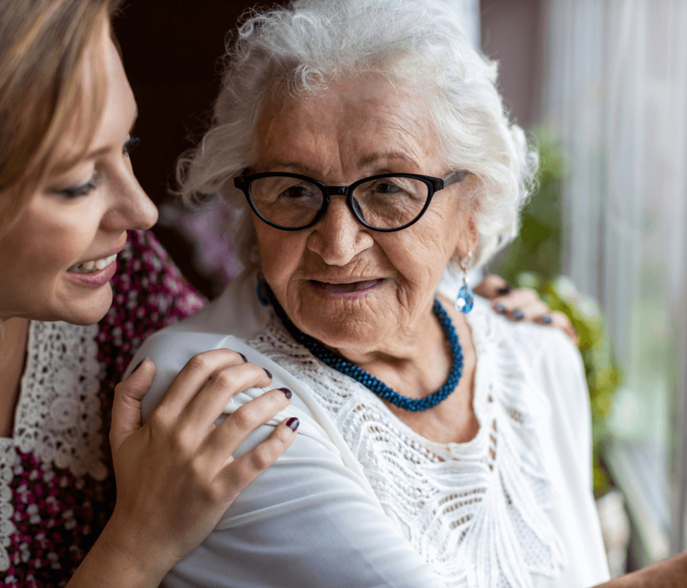 A smiling elderly woman with glasses is embraced by a younger woman, both looking happy near a window. - Home Instead