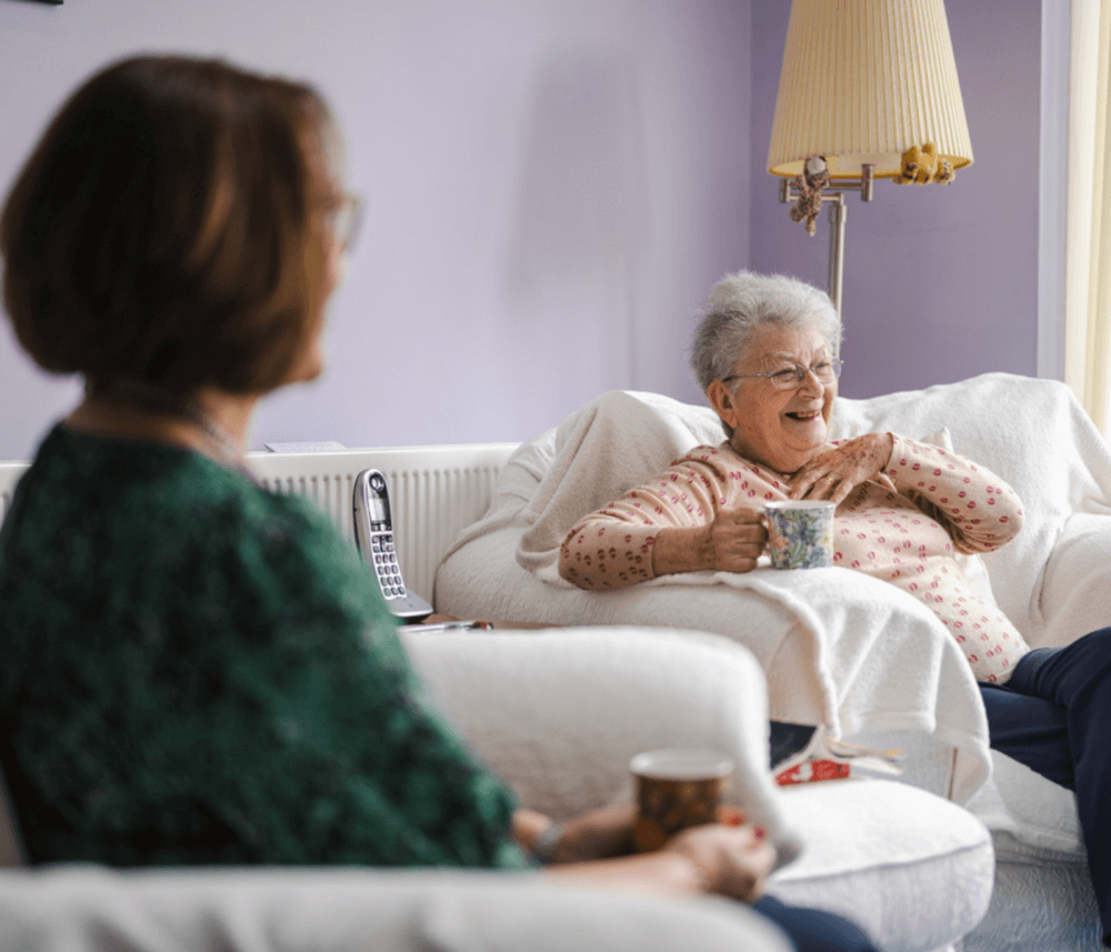 Two women sitting and chatting, one elderly woman laughing with a mug in hand, the other blurred in the foreground. - Home Instead