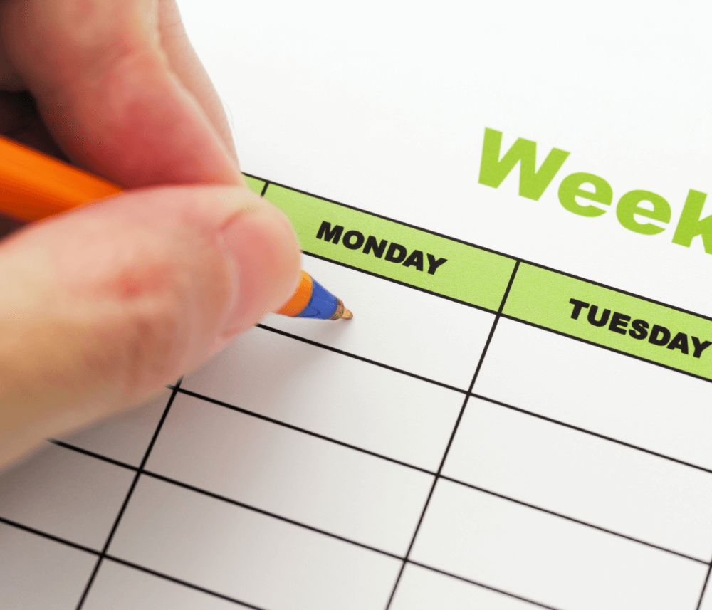 A close-up of a hand holding a pen, writing on a weekly calendar with the days of the week labeled, highlighting Monday. - Home Instead