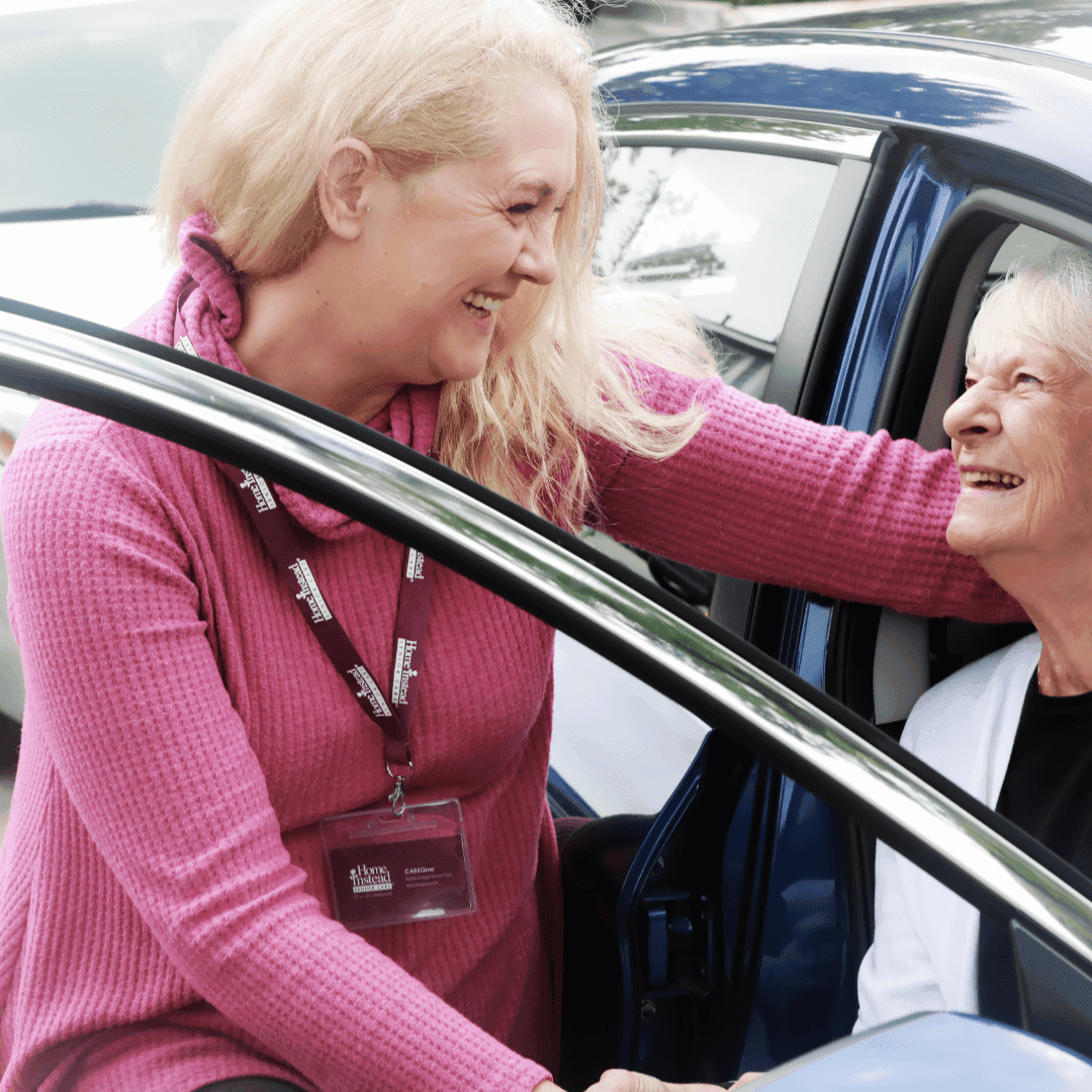 A woman in a pink sweater helps another woman with gray hair out of a car while they both smile warmly at each other. - Home Instead
