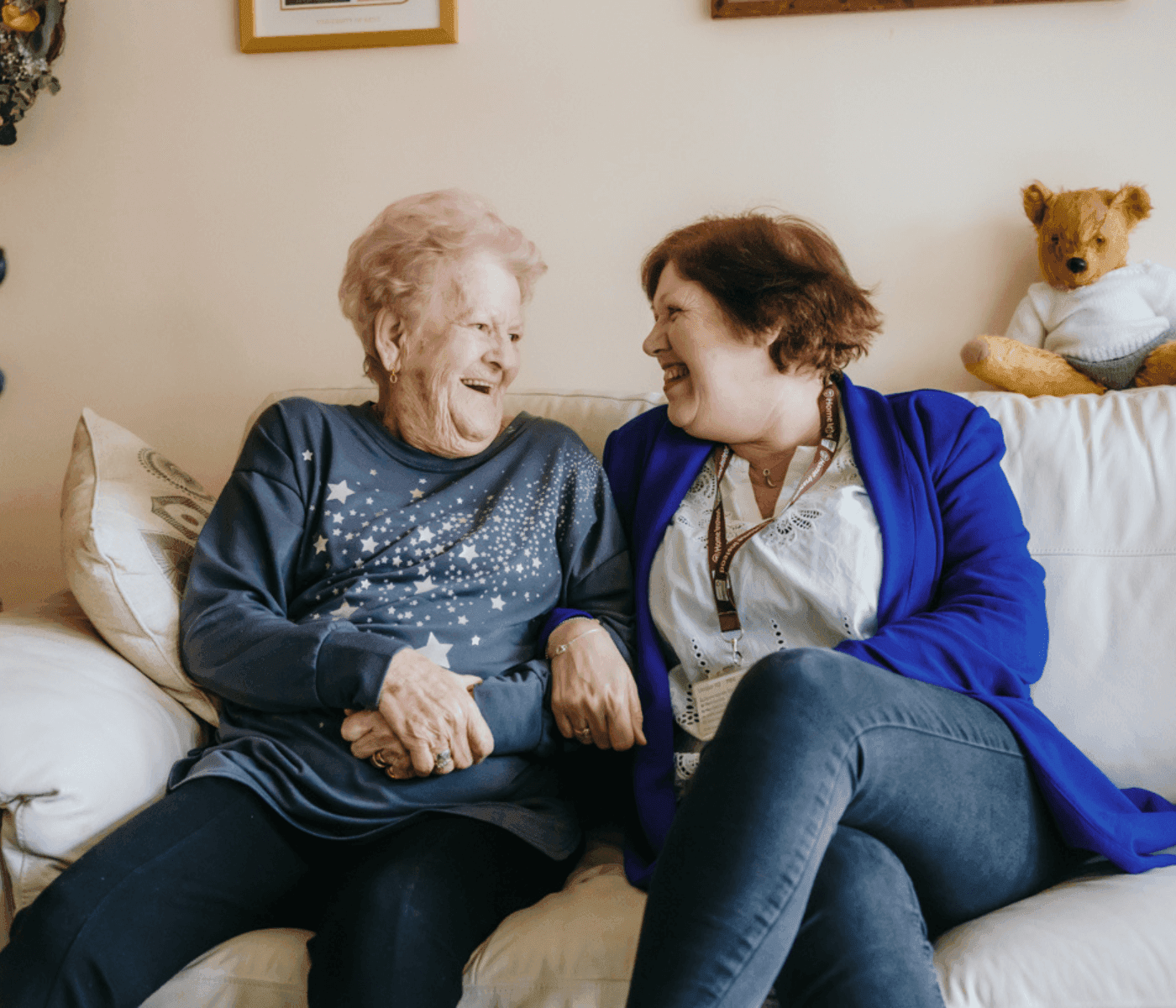 Two women, an elderly woman with pink hair and a younger woman with short brown hair, laugh together on a couch. - Home Instead