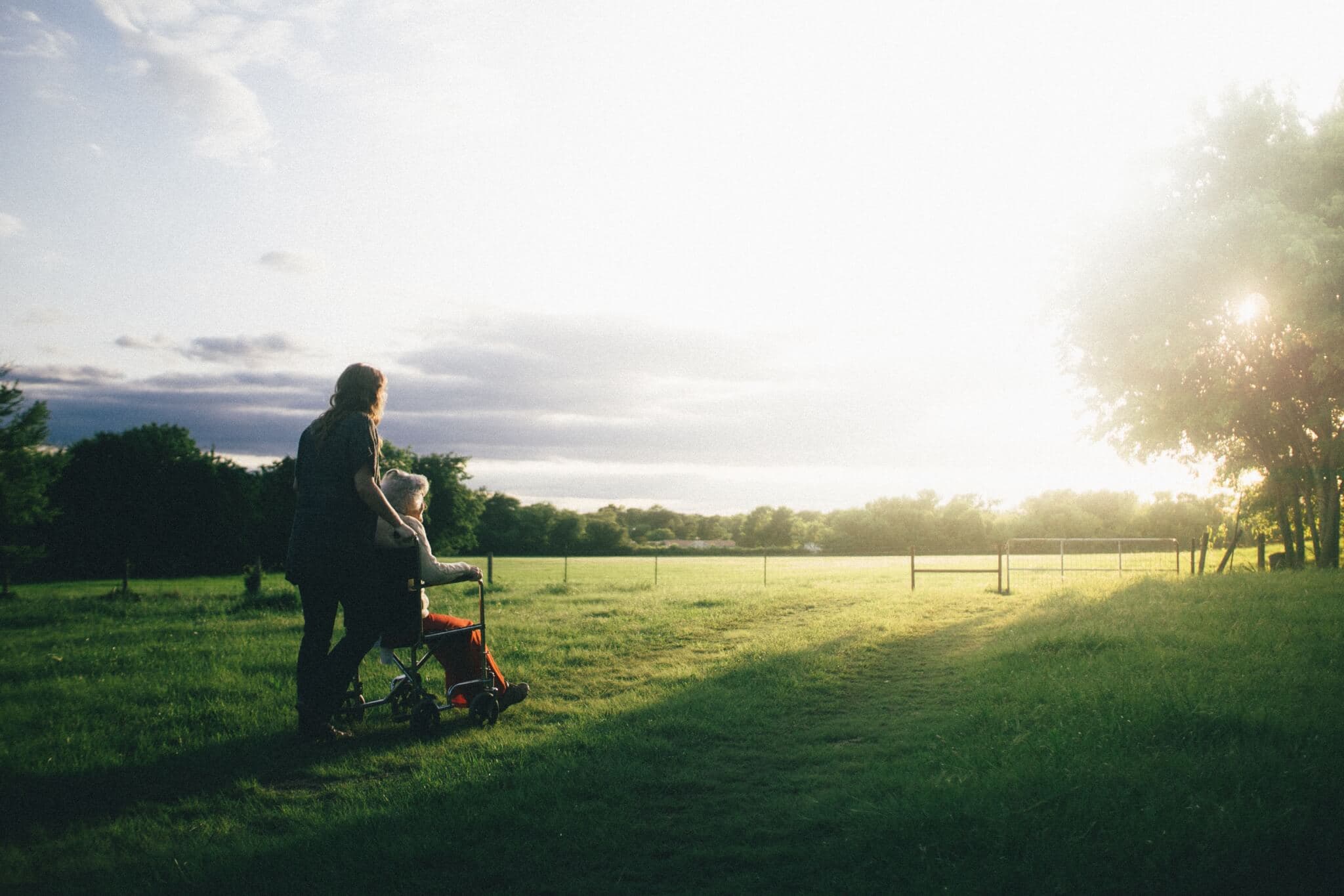 A person pushes another in a wheelchair along a grassy path, with the sun setting over the horizon in a rural area. - Home Instead