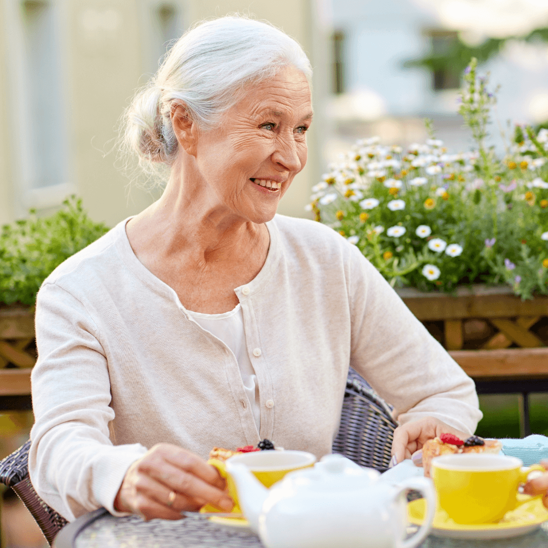 Elderly woman with gray hair smiles while enjoying tea and dessert at an outdoor table with flowers in the background. - Home Instead