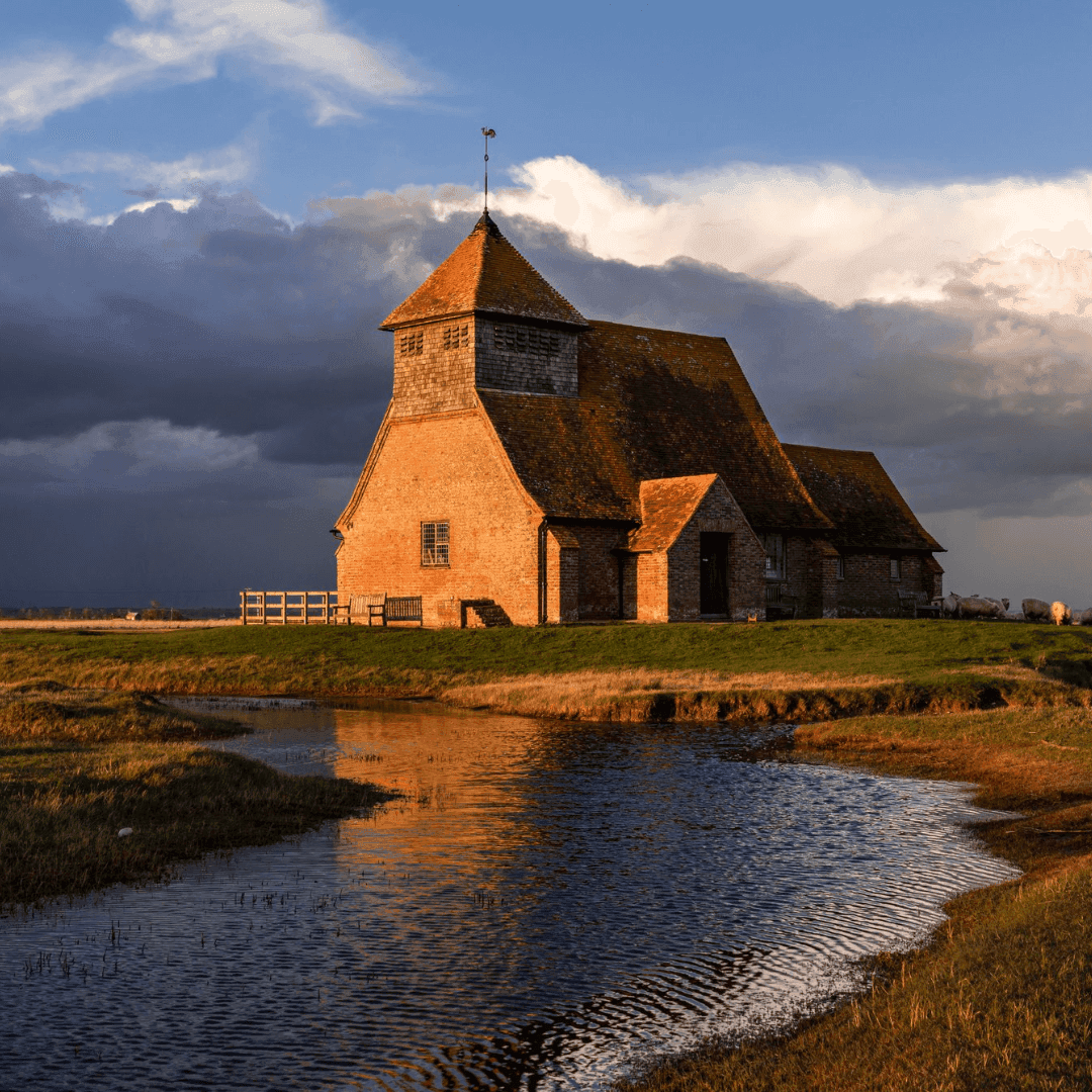 A lone, rustic brick church sits by a reflective stream under a dramatic cloudy sky during golden hour. - Home Instead