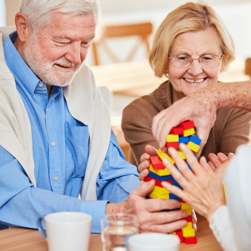 Two elderly people smiling and building a tower with colorful toy blocks on a table, assisted by a third person. - Home Instead