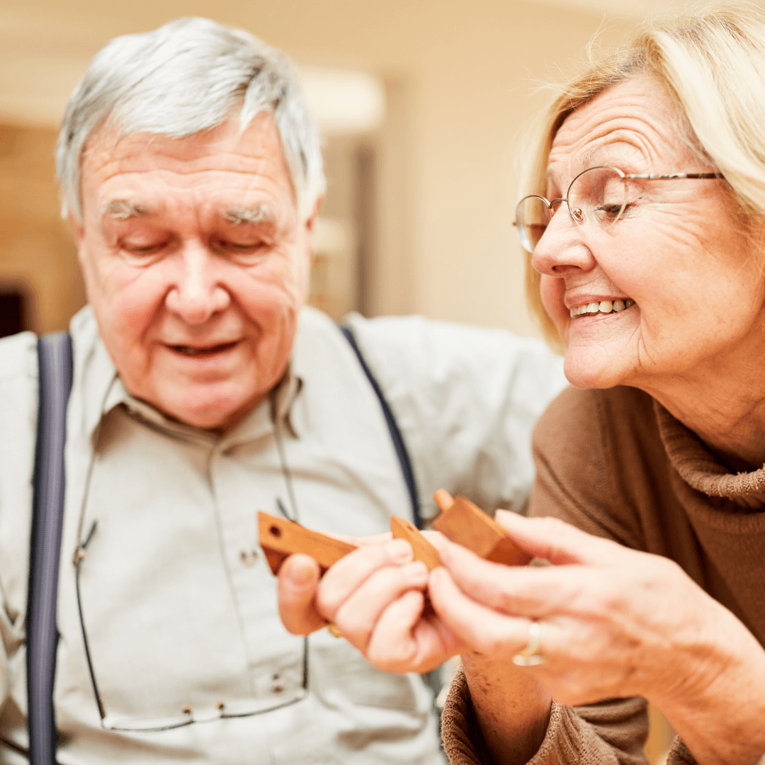 An elderly couple smiling and looking at each other while holding wooden blocks inside a house. - Home Instead