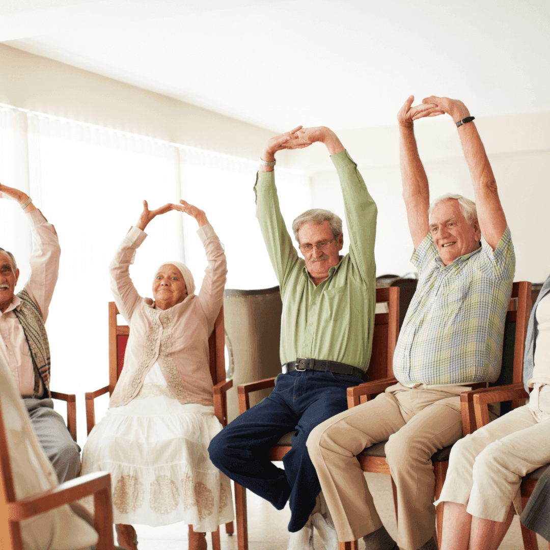 A group of elderly people sit on chairs in a bright room, engaging in light exercises with arms raised overhead. - Home Instead