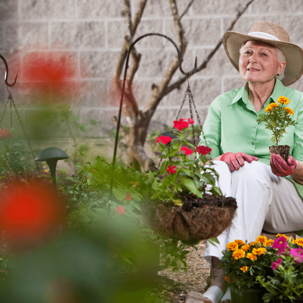 Elderly woman in garden wearing a hat, holding a potted plant, seated among colorful flowers and greenery. - Home Instead