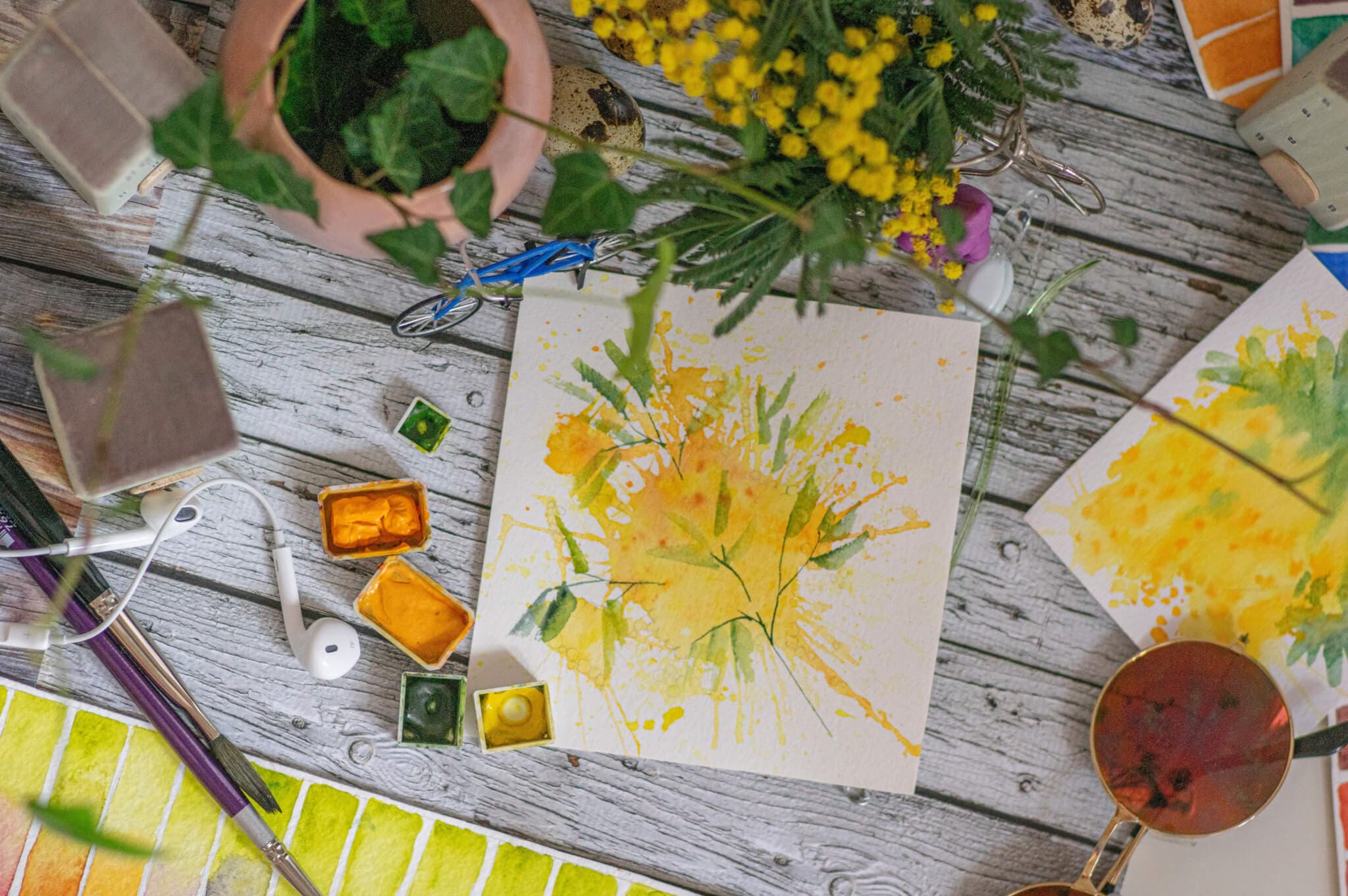 A bright watercolor painting on a wooden surface surrounded by painting tools, greenery, and yellow flowers. - Home Instead