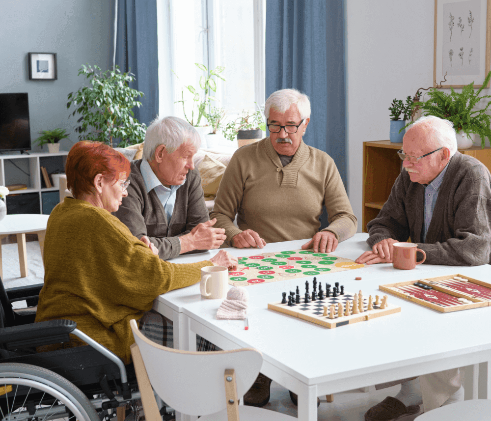 Four elderly people playing a board game at a table, with a chess set nearby and mugs on the table. - Home Instead