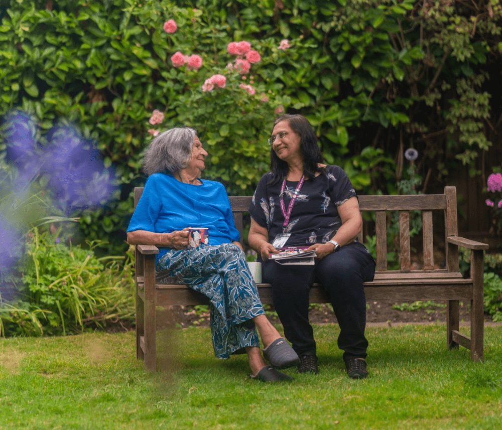 Two women sit and smile at each other on a garden bench surrounded by greenery and blooming flowers. - Home Instead