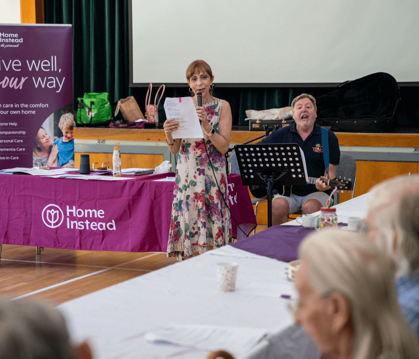 A woman holding a microphone and a man playing guitar host an event at a hall with a "Home Instead" table and banner. - Home Instead