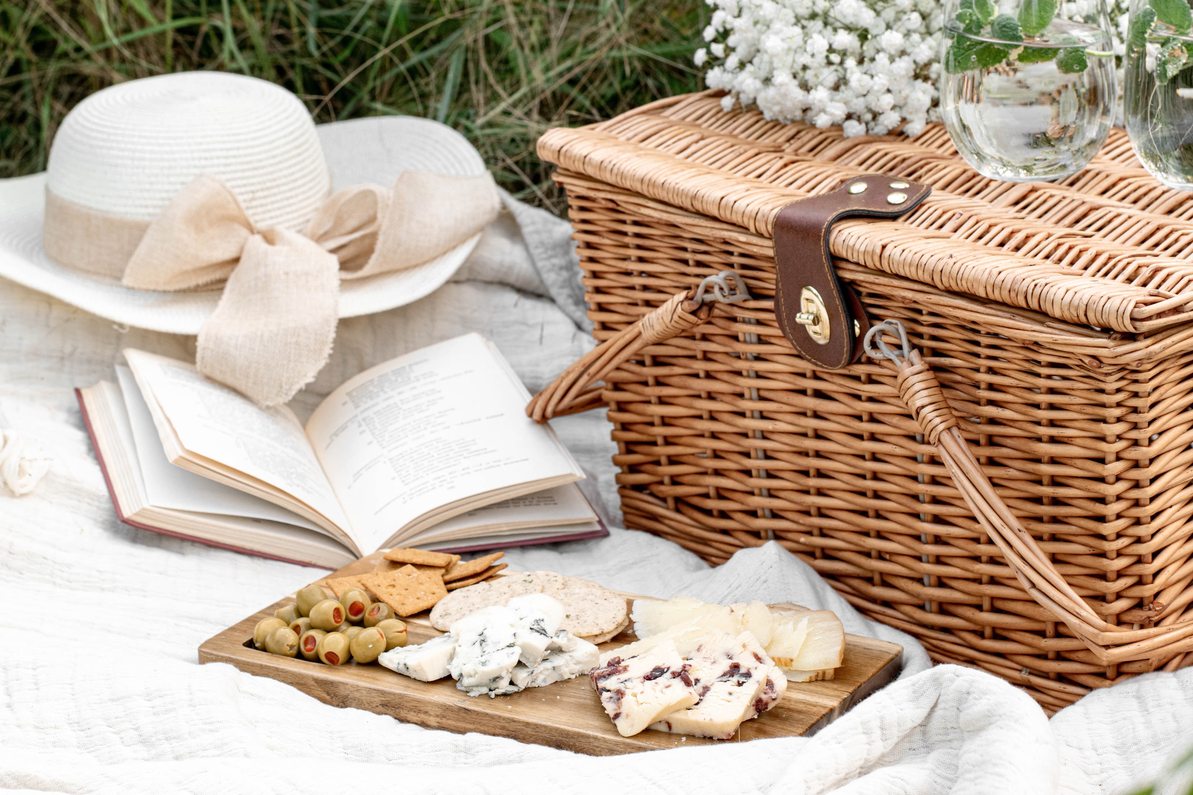 A picnic setup with a wicker basket, open book, white hat, and cheese platter on a white blanket in the grass. - Home Instead