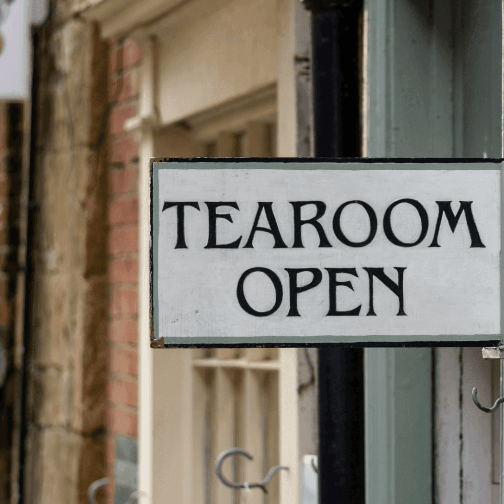 Sign reading "Tearoom Open" hanging outside a shop door on a street with brick and wooden buildings. - Home Instead