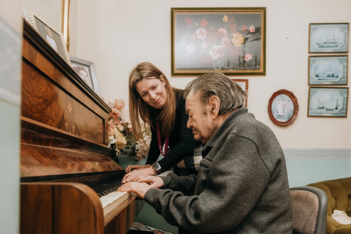 An elderly man plays piano assisted by a woman in a room with floral and nautical paintings. - Home Instead