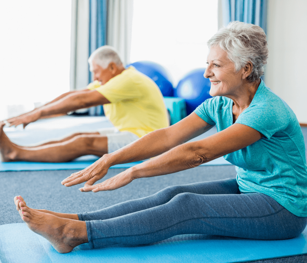 Two older adults in workout clothes perform seated forward bends on yoga mats, smiling in a bright room. - Home Instead