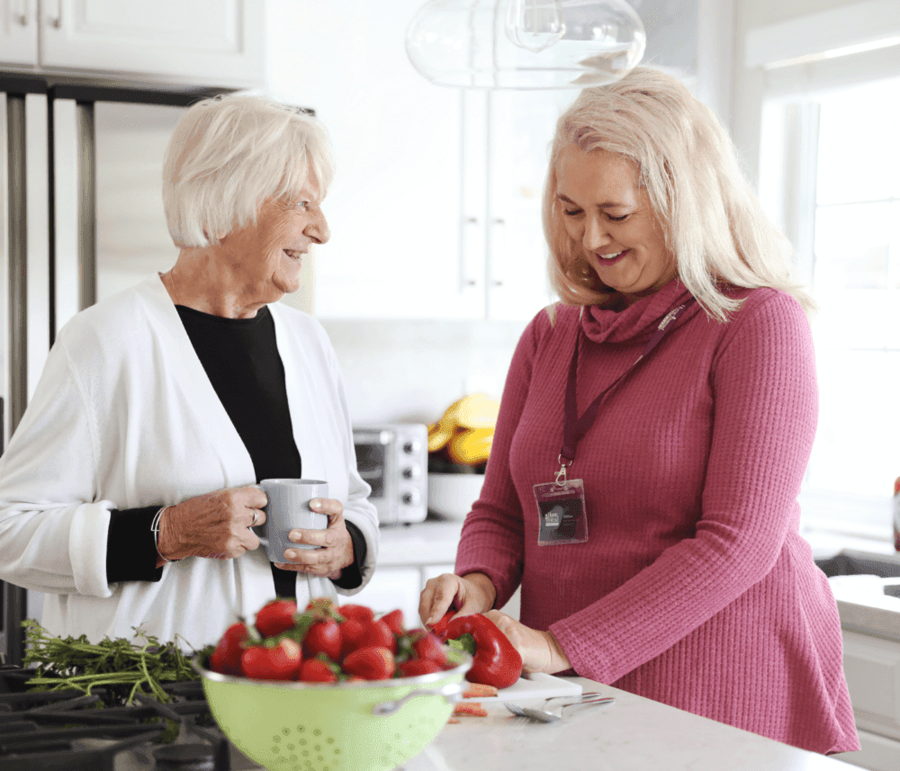 Two women in a kitchen, one chopping vegetables and the other holding a mug, with a colander of strawberries in front. - Home Instead