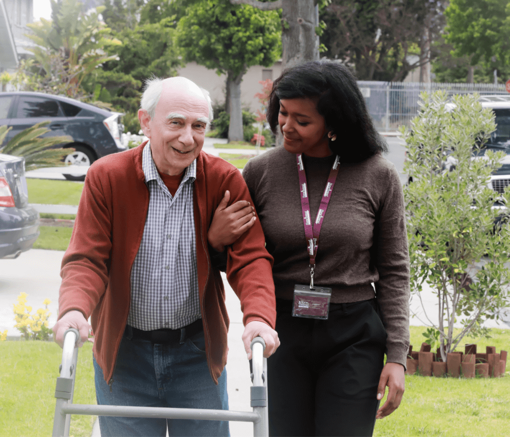 A senior man using a walker is assisted by a caregiver. They are walking together outside in a residential area. - Home Instead
