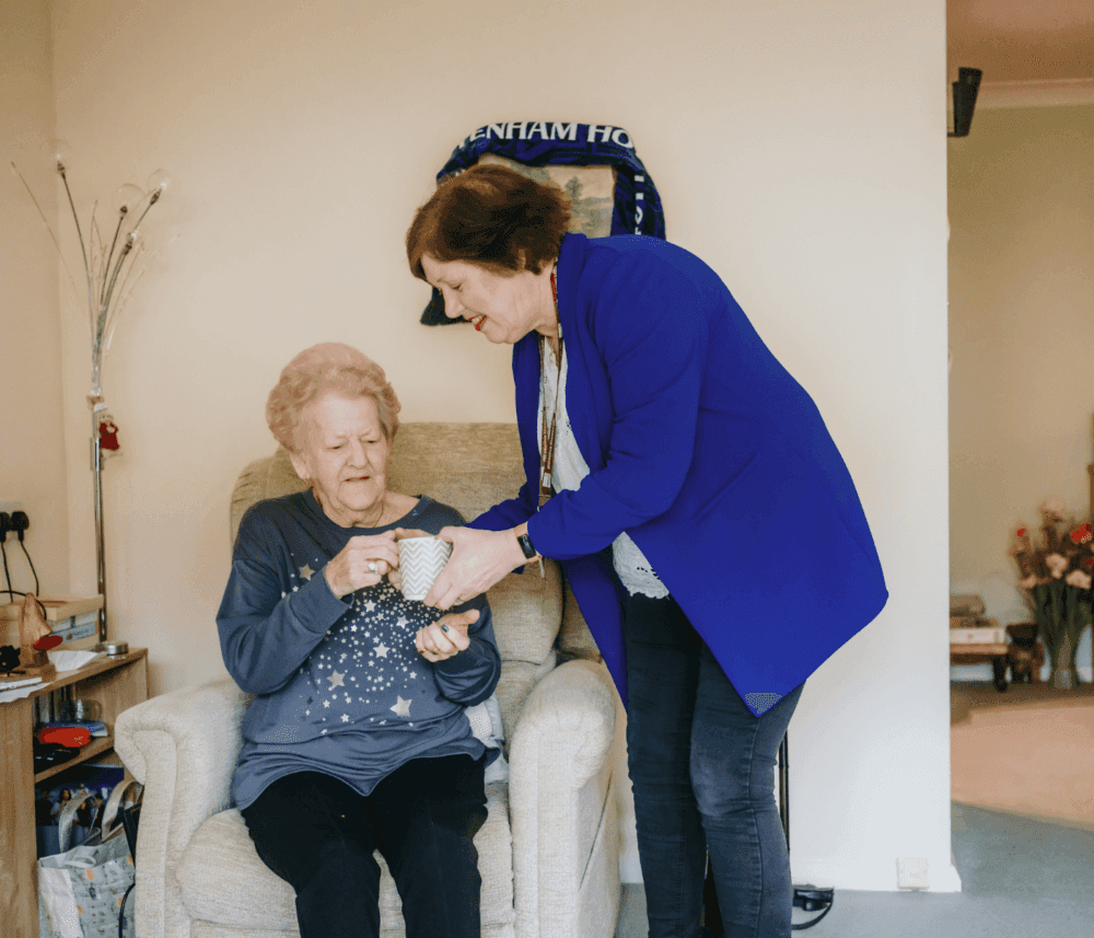 A woman in a blue jacket hands a mug to an elderly woman seated in a chair in a cozy room. - Home Instead