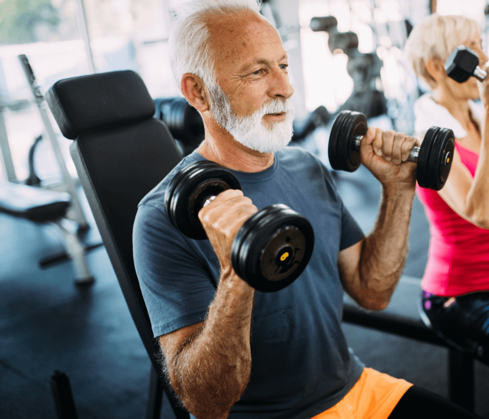 An elderly man with gray hair exercises with dumbbells in a gym, alongside a woman in a pink top in the background. - Home Instead