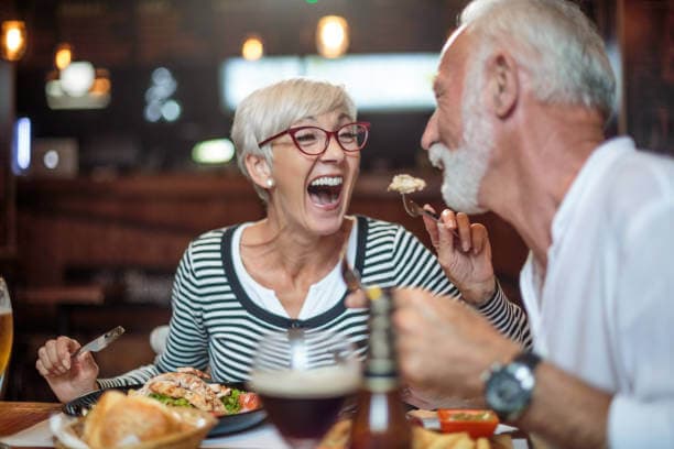 An older couple laughing and enjoying a meal together at a restaurant. - Home Instead