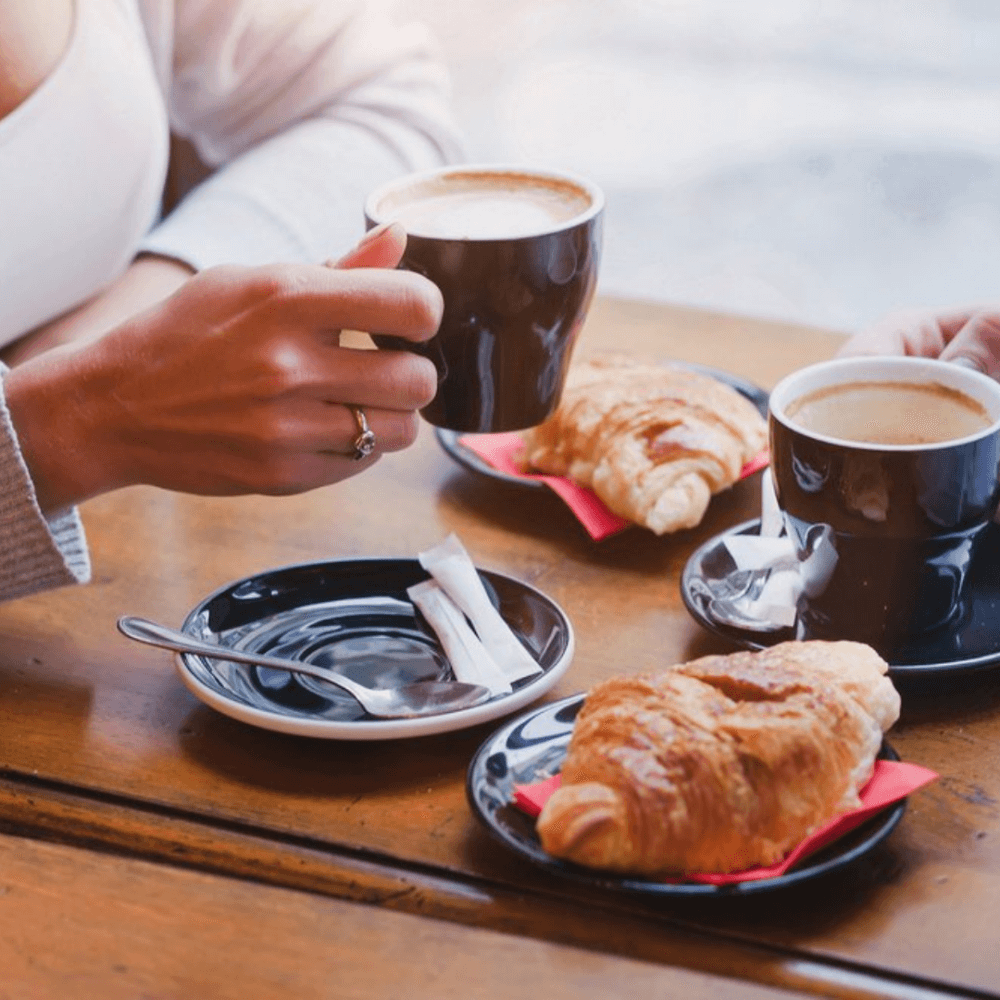 Two people enjoying coffee and croissants at a wooden table. - Home Instead