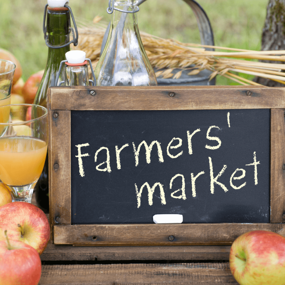 Chalkboard sign reading "Farmers' market" surrounded by apples, various bottles, and a bundle of wheat. - Home Instead