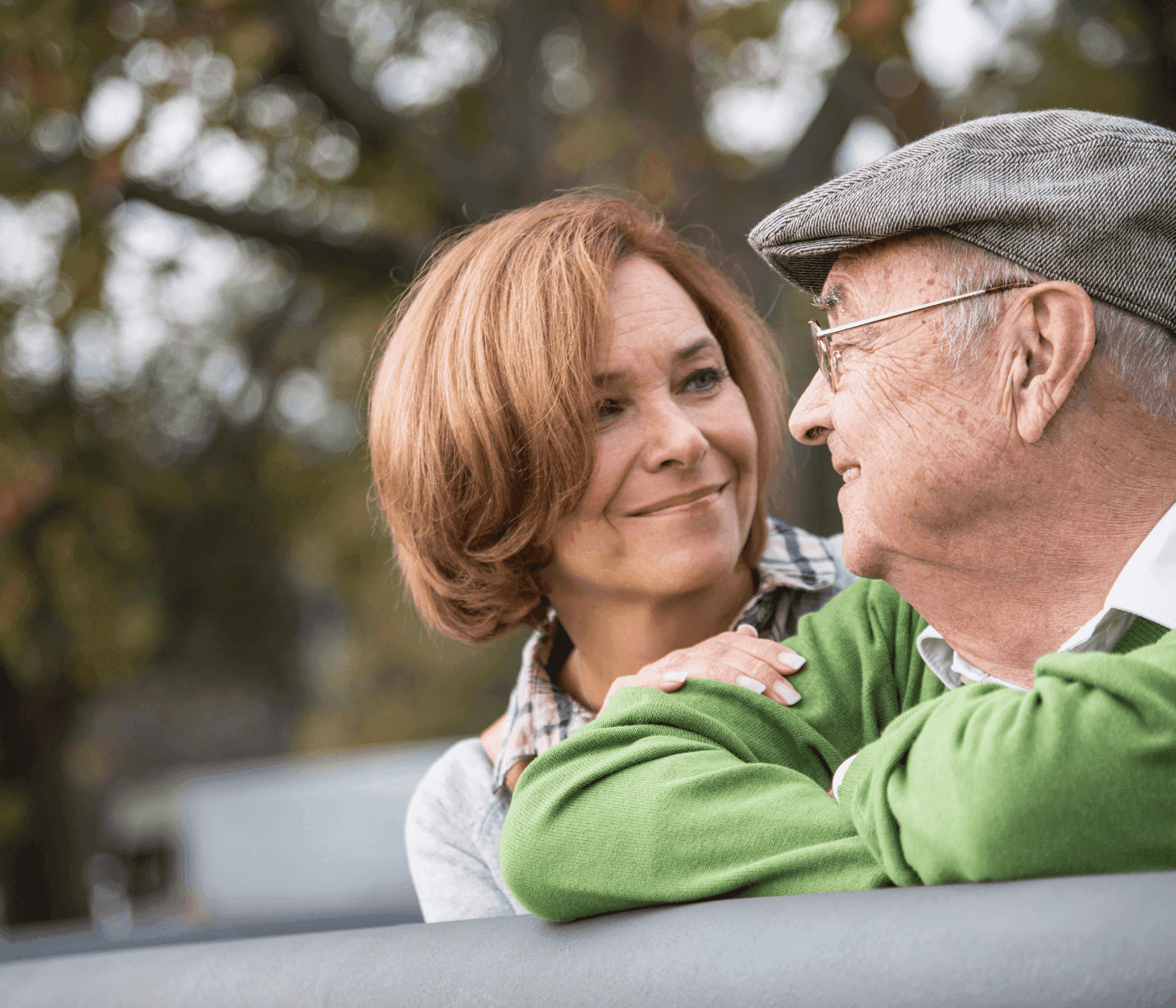 An older man and a middle-aged woman smile at each other warmly while leaning on a fence in a park. - Home Instead
