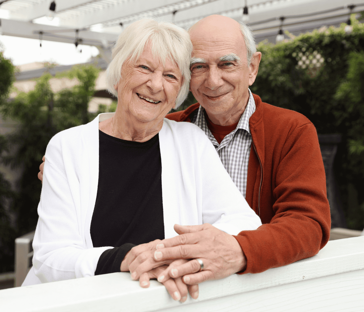 An elderly couple smiling, standing closely together outdoors, with greenery in the background. - Home Instead