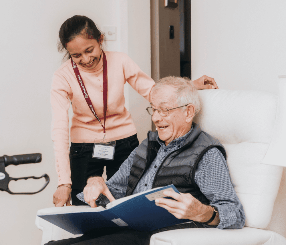 Caregiver helping an elderly man read a book while they both smile in a cozy living room setting. - Home Instead