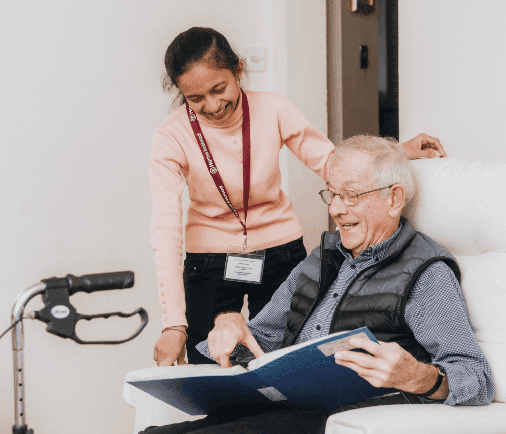A caregiver and an elderly man happily look at a photo album together in a cozy indoor setting. - Home Instead