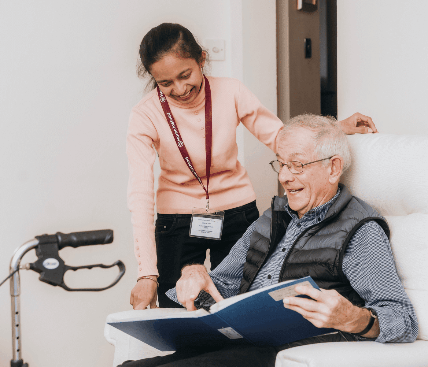 A caregiver and an elderly man happily look at a photo album together in a cozy indoor setting. - Home Instead