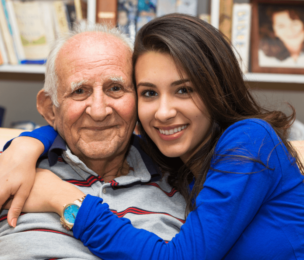 A smiling young woman in a blue shirt hugs an elderly man in a striped shirt, both seated and happy. - Home Instead