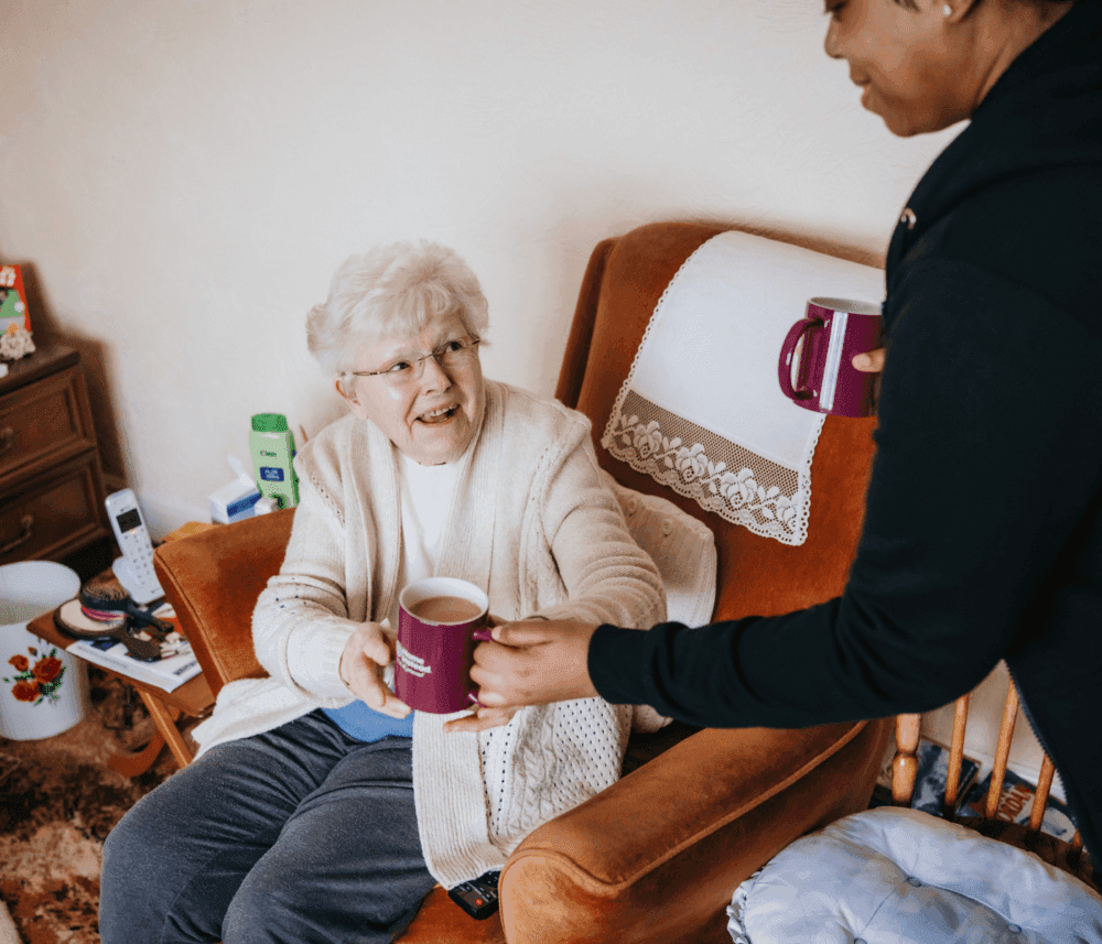 An elderly woman sitting in a chair smiles as she receives a mug of tea from a caregiver standing next to her. - Home Instead