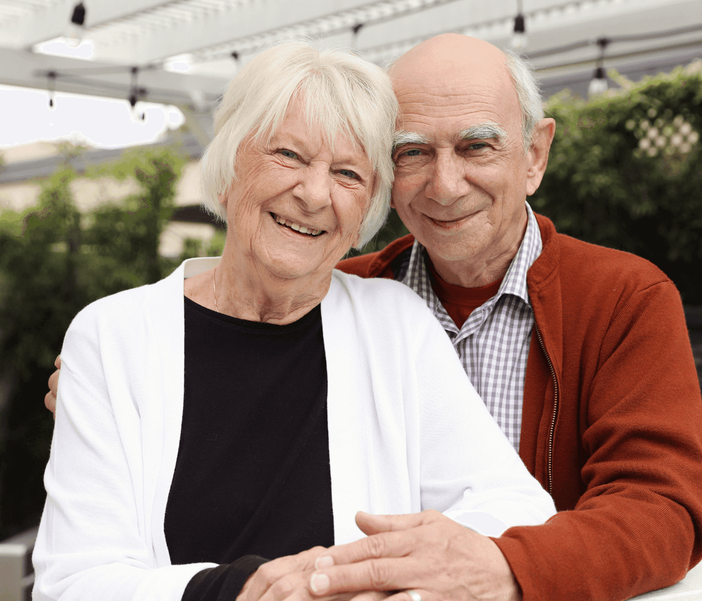 An elderly couple smiles warmly at the camera, embracing each other outdoors under a pergola. - Home Instead