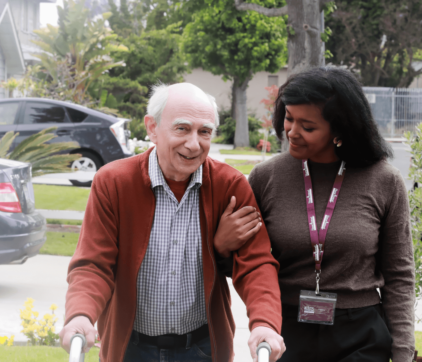 An older man with a walker smiles, assisted by a woman who holds his arm. They are outdoors on a sunny day. - Home Instead