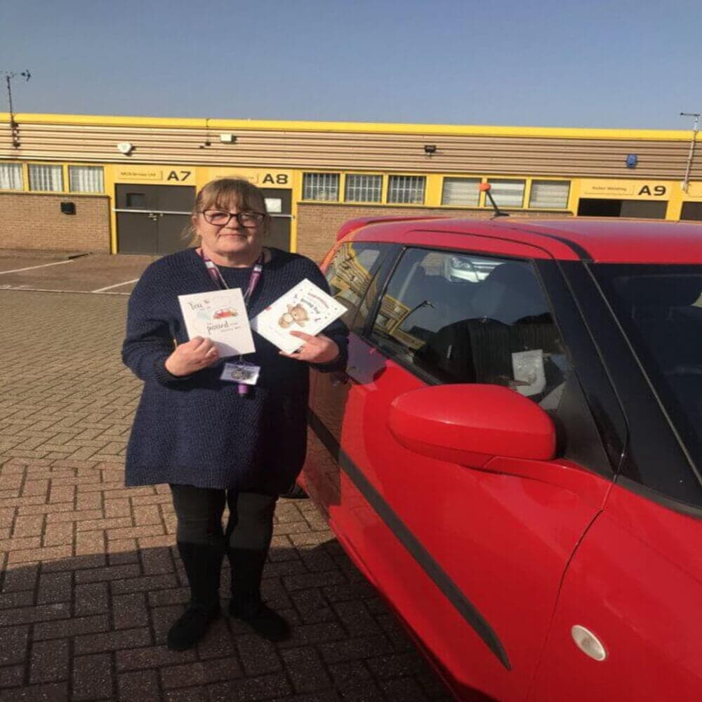 A woman stands beside a red car, holding greeting cards and smiling in a parking lot with yellow industrial buildings. - Home Instead