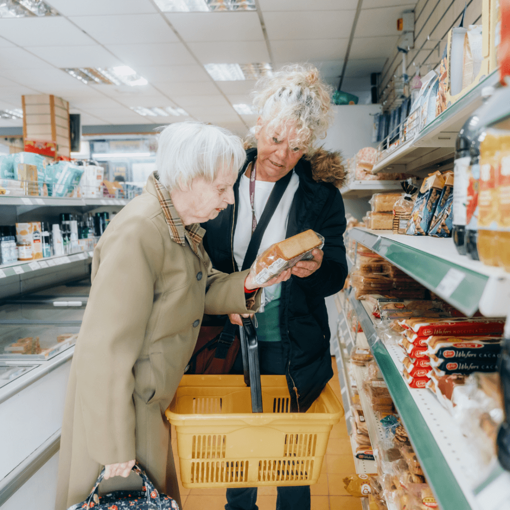 Two elderly people in a grocery store aisle, one holding bread and the other with a shopping basket, examining product. - Home Instead
