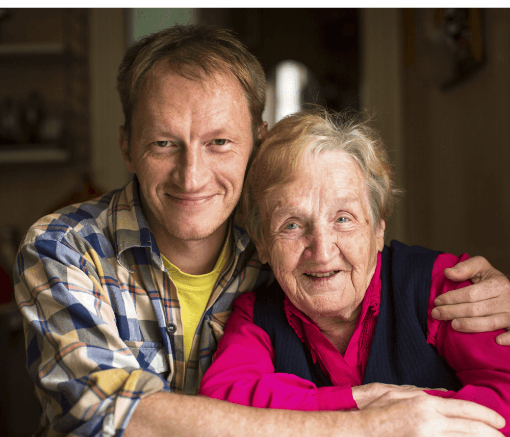 A smiling man in a plaid shirt and an older woman in a pink blouse pose together, with arms around each other. - Home Instead