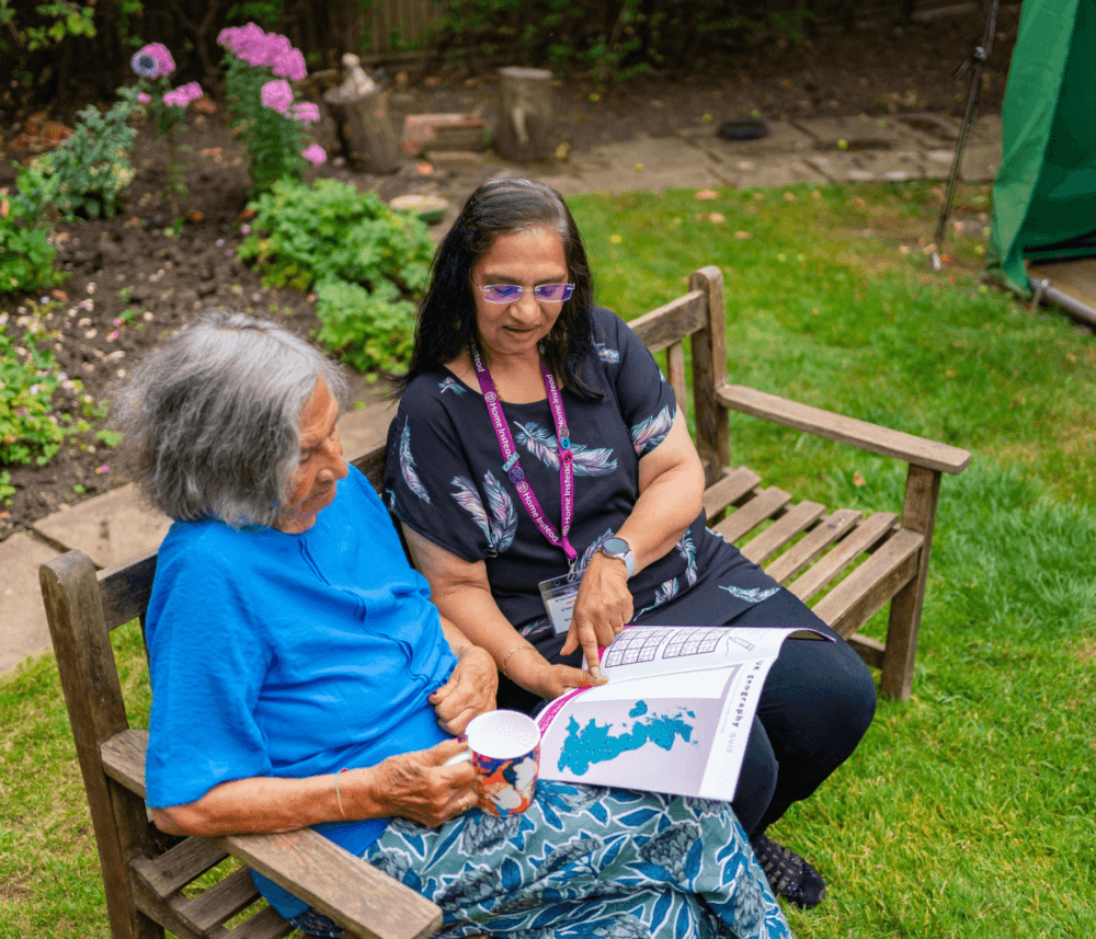 Two women sitting on a wooden bench in a garden, looking at a map and enjoying a conversation. - Home Instead