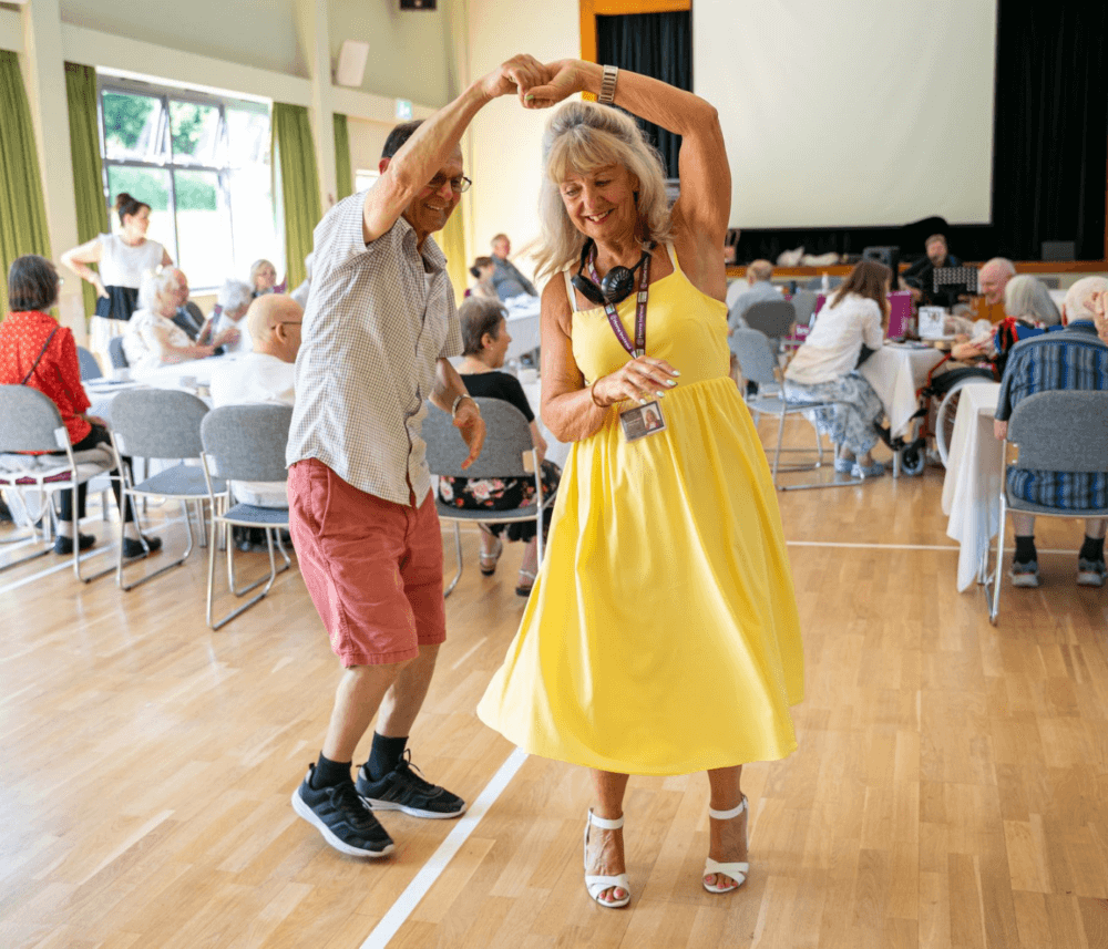 An elderly man and woman dance joyfully in a community center while others sit around tables in the background. - Home Instead